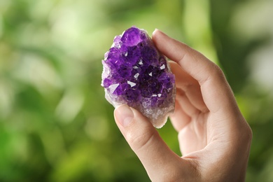Woman holding beautiful purple amethyst gemstone on blurred green background, closeup