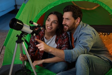Happy couple using telescope to look at stars while sitting in camping tent indoors