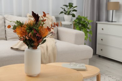 Photo of Vase with bouquet of beautiful leucospermum flowers, book and glasses on wooden nesting tables near beige sofa in room, space for text