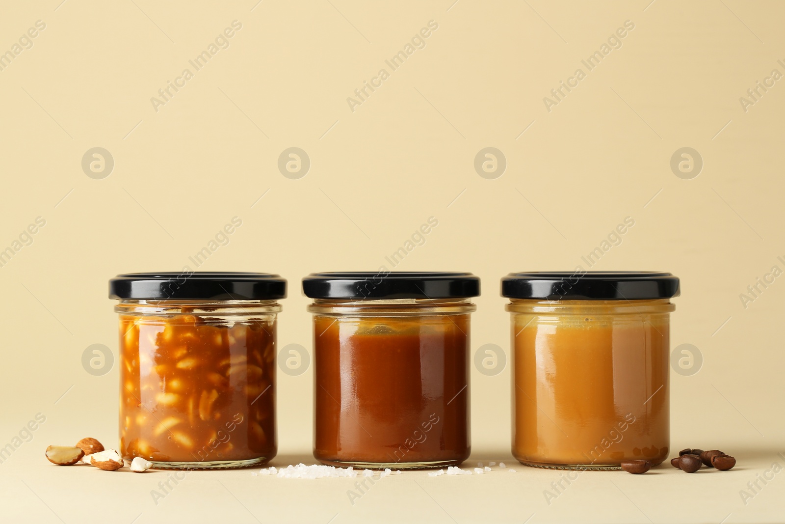 Photo of Tasty salted caramel in jars with peanuts, coffee beans and salt on pale yellow background