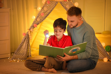 Photo of Father and son with flashlight reading book at home