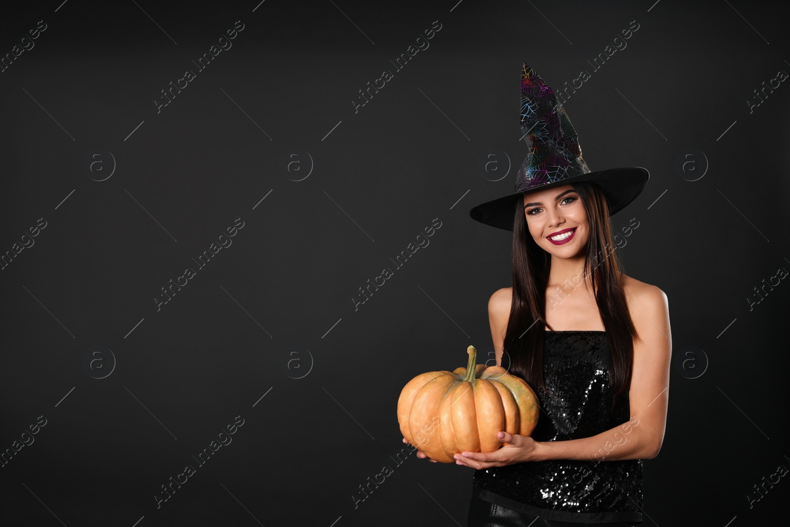 Photo of Beautiful woman wearing witch costume with pumpkin for Halloween party on black background, space for text