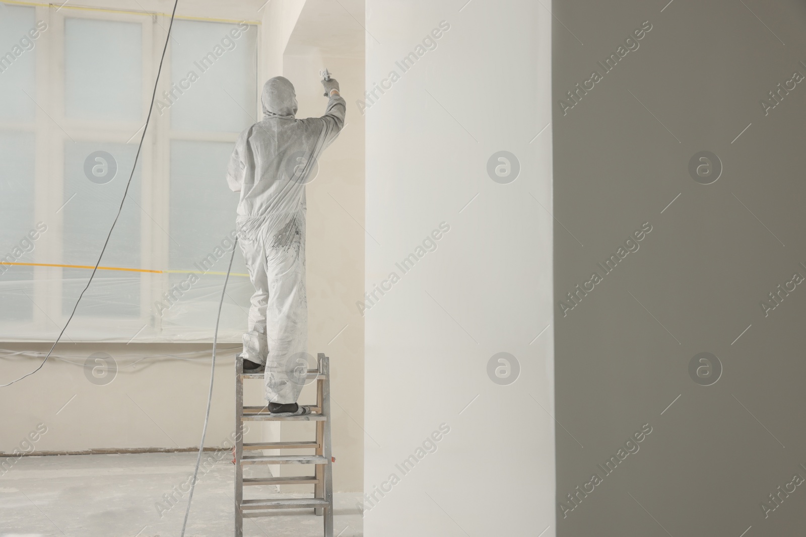 Photo of Decorator in protective overalls painting wall with spray gun indoors