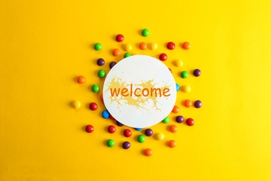 Image of Colorful candies and card with word WELCOME on yellow background, flat lay