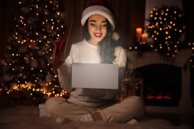 Young woman wearing Santa hat opening Christmas gift on floor at home