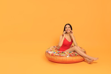 Photo of Happy young woman with beautiful suntan talking by phone on inflatable ring against orange background, space for text