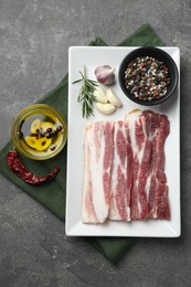Photo of Pieces of raw pork belly, oil, peppercorns, rosemary and garlic on grey textured table, flat lay