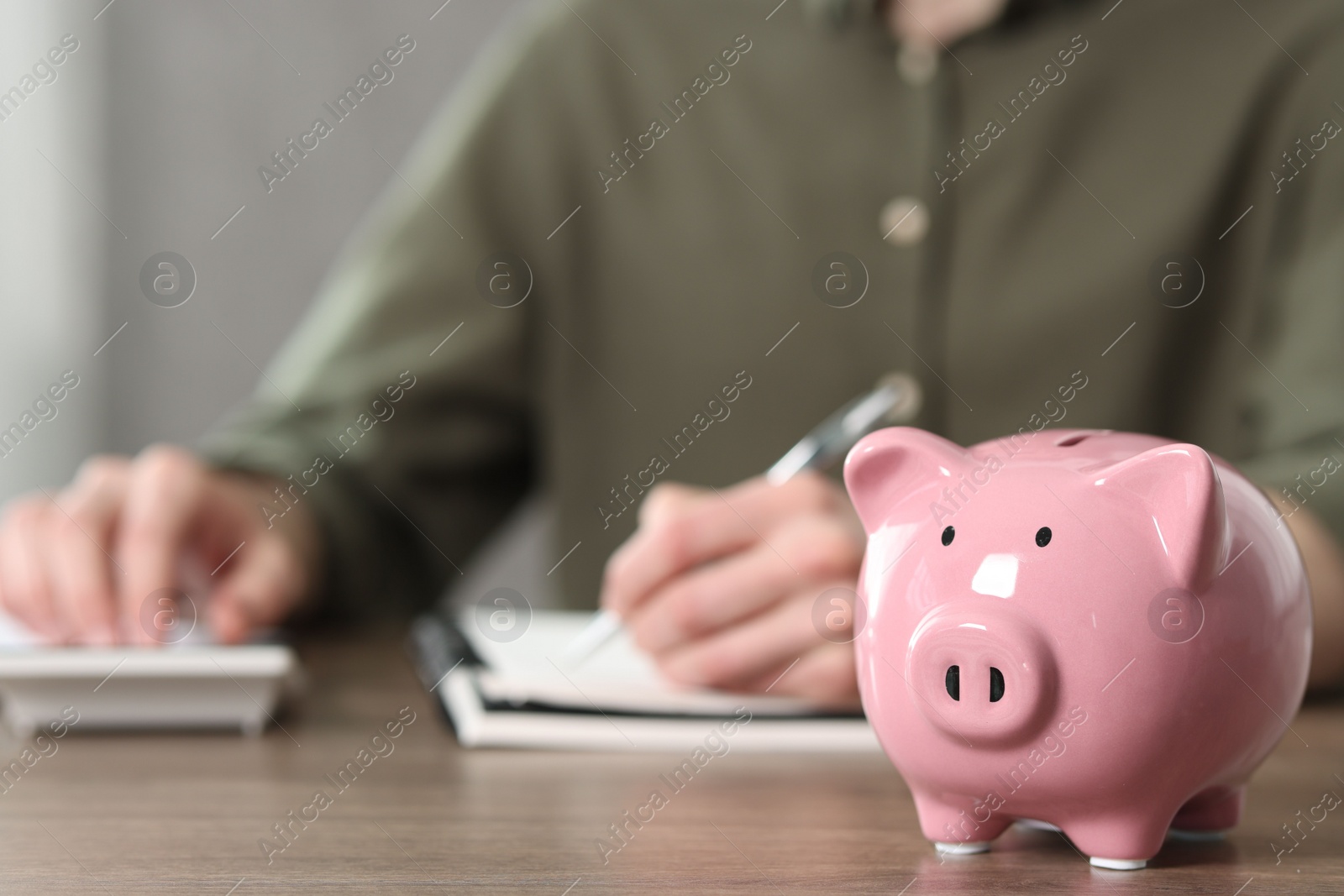 Photo of Financial savings. Man writing down notes and using calculator at wooden table, focus on piggy bank