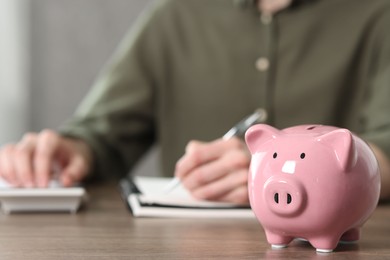 Financial savings. Man writing down notes and using calculator at wooden table, focus on piggy bank