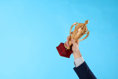 Photo of Businessman holding gold trophy cup on light blue background, closeup. Space for text