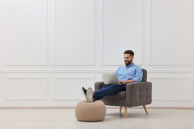 Handsome man using laptop while sitting in armchair near white wall indoors, space for text