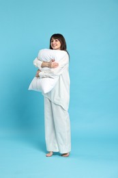 Happy woman in pyjama holding pillow on light blue background