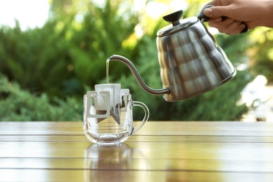 Photo of Woman pouring hot water into glass cup with drip coffee bag from kettle at wooden table, closeup