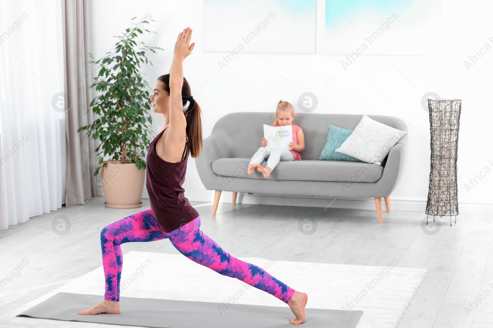 Photo of Woman doing fitness exercises while her daughter sitting on sofa at home