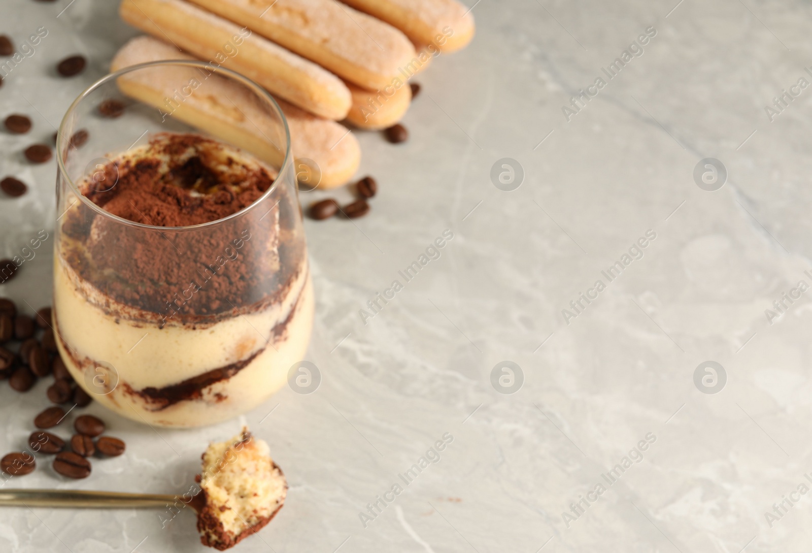 Photo of Tasty tiramisu in glass, coffee beans, spoon and biscuits on light grey table, closeup. Space for text