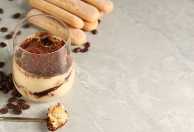 Photo of Tasty tiramisu in glass, coffee beans, spoon and biscuits on light grey table, closeup. Space for text