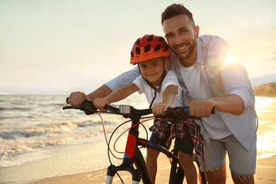 Happy father teaching son to ride bicycle on sandy beach near sea at sunset
