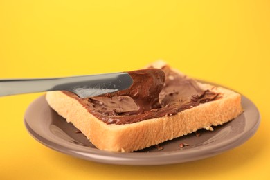 Photo of Spreading chocolate paste onto bread on yellow background, closeup