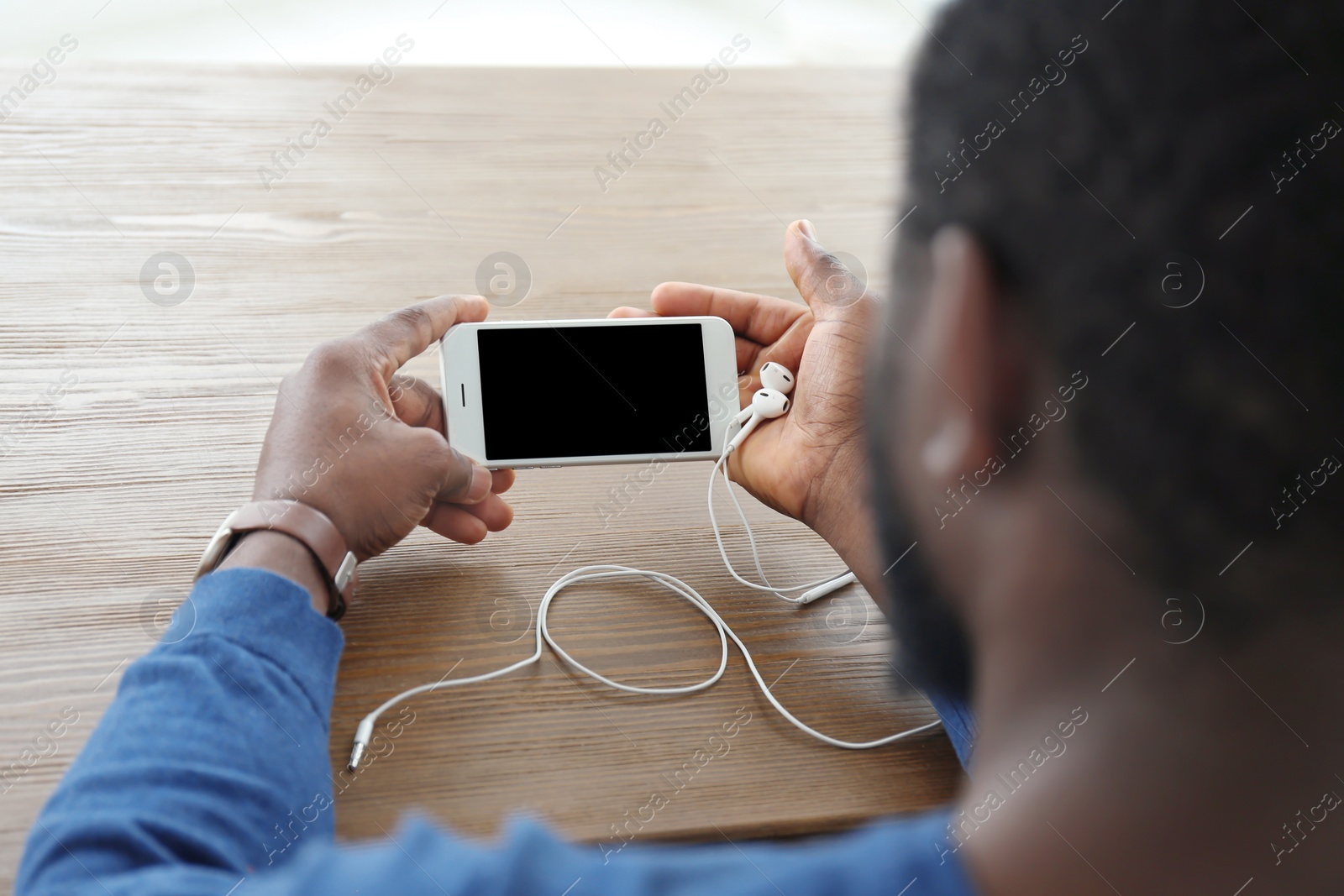 Photo of African-American man holding mobile phone with blank screen in hands at table indoors