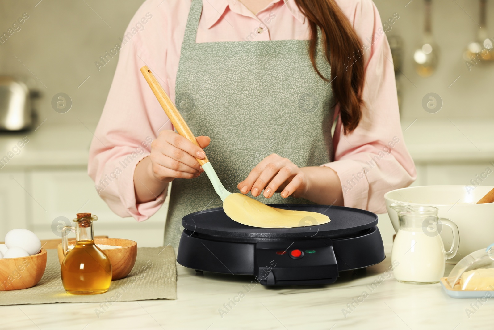 Photo of Woman cooking delicious crepe on electric maker at white marble table in kitchen, closeup