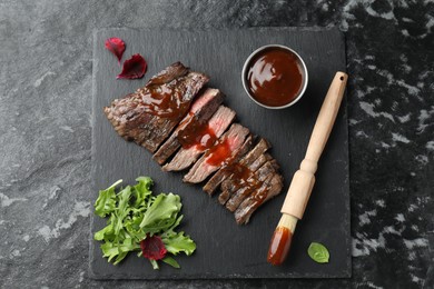 Photo of Pieces of delicious roasted beef meat with sauce and greens on black table, top view