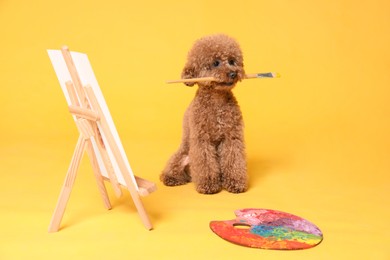 Photo of Dog artist. Cute Maltipoo holding brush near easel with canvas and palette on orange background