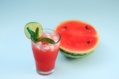 Photo of Glass of delicious drink with lime, ice cubes and cut fresh watermelon on light blue background