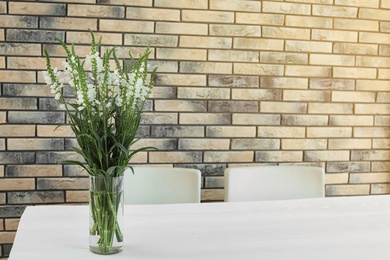 Photo of Vase with beautiful flowers on white table against brick wall. Stylish interior