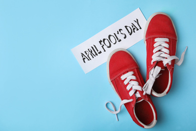 Photo of Sport shoes tied together and note with phrase APRIL FOOL'S DAY on light blue background, flat lay. Space for text