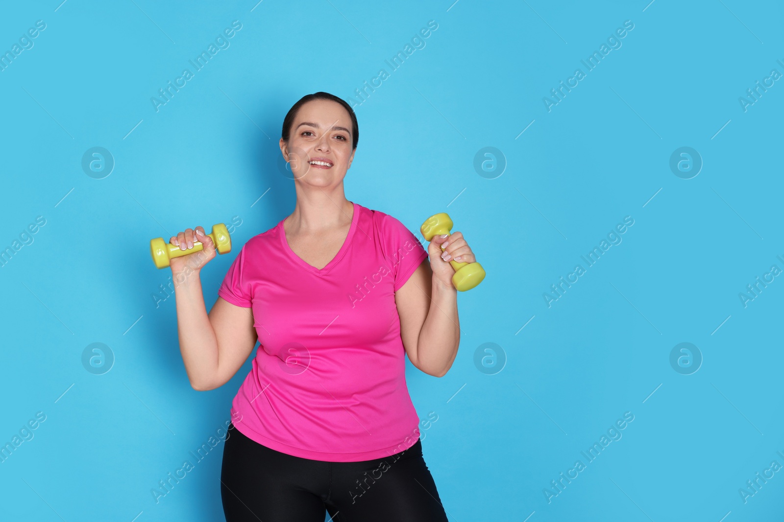Photo of Happy overweight woman doing exercise with dumbbells on light blue background, space for text