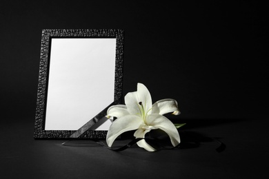 Photo of Funeral photo frame with ribbon and white lily on black background. Space for design