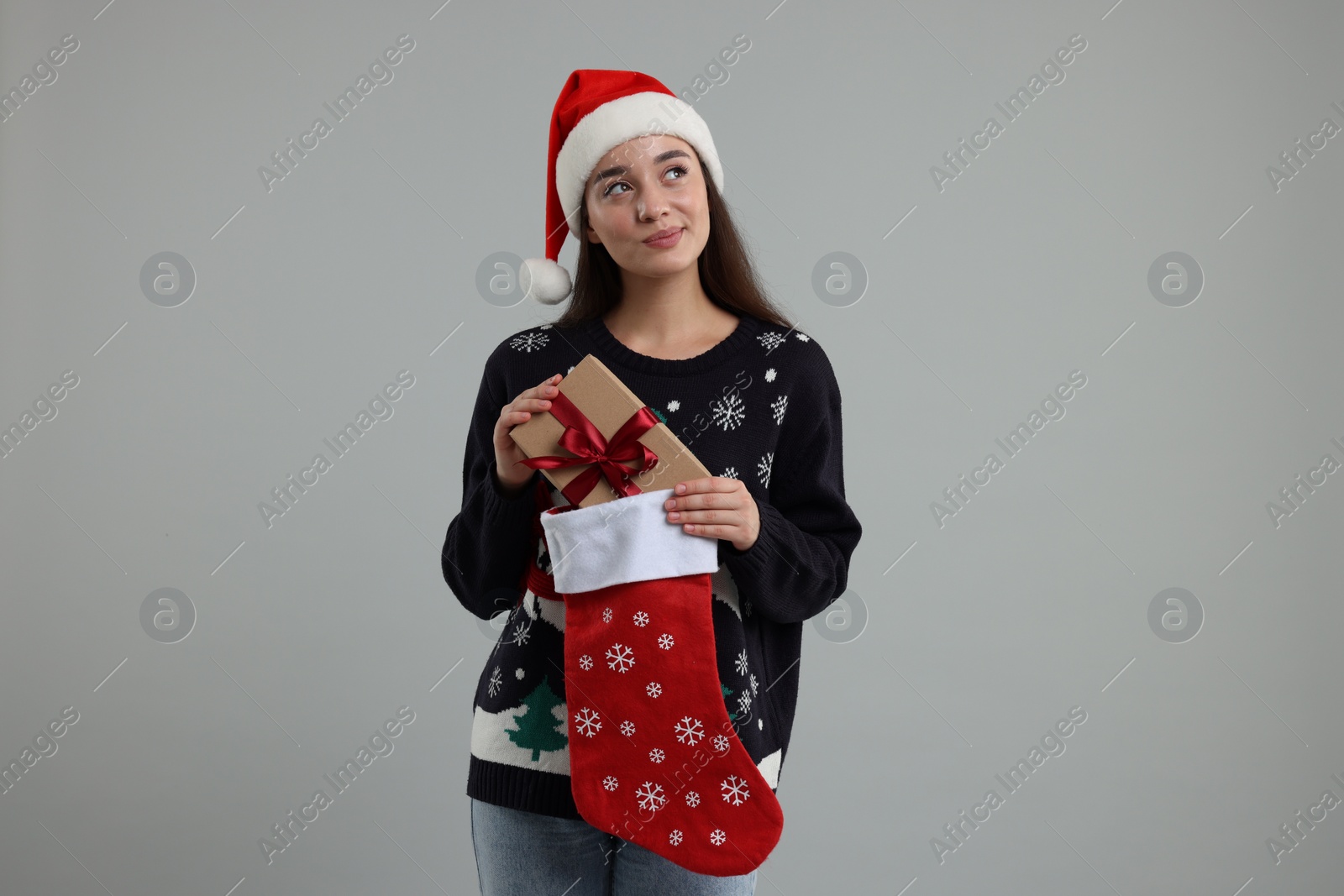 Photo of Young woman in Christmas sweater and Santa hat taking gift from stocking on grey background