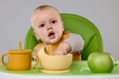 Cute little baby eating healthy food in high chair on gray background