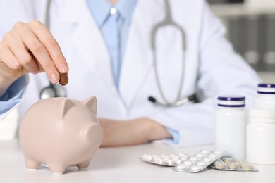 Doctor putting coin into piggy bank at white table indoors, closeup