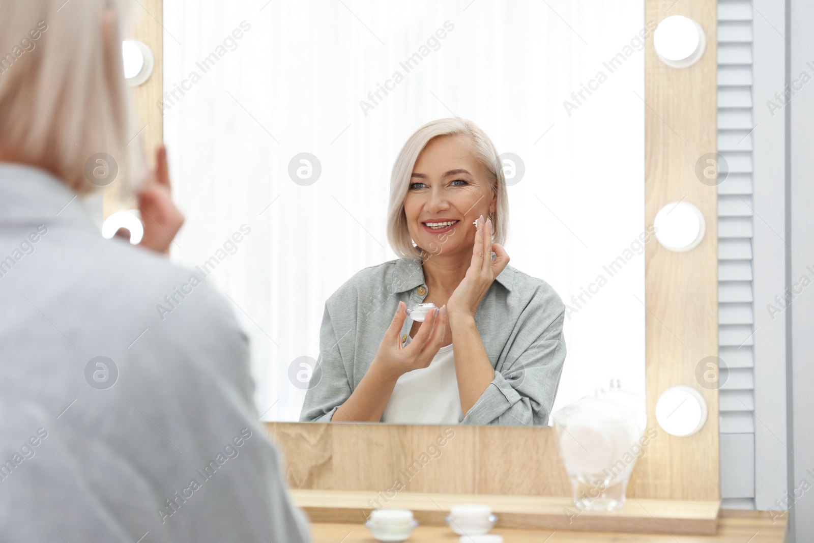 Photo of Portrait of charming mature woman with healthy beautiful face skin and natural makeup applying cream indoors