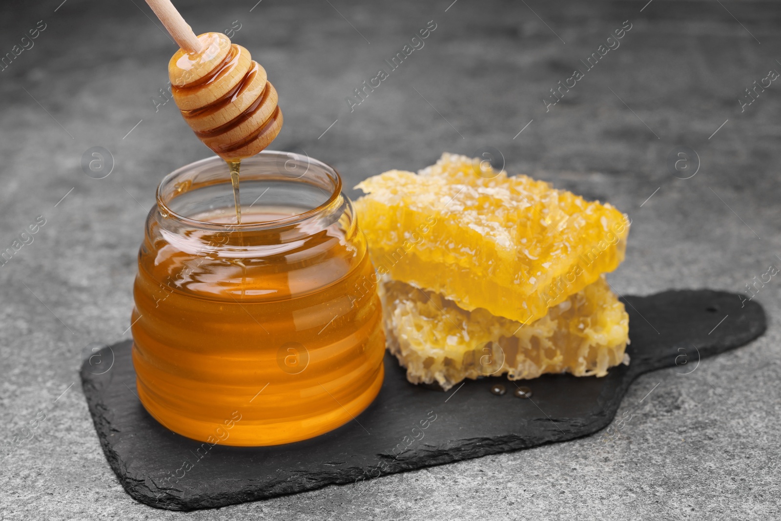Photo of Pouring sweet golden honey from dipper into jar and pieces of honeycomb on grey textured table
