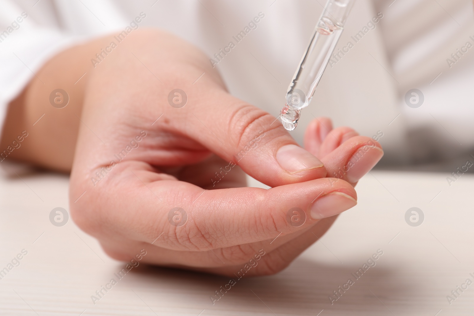 Photo of Woman applying cosmetic serum onto finger at white table, closeup