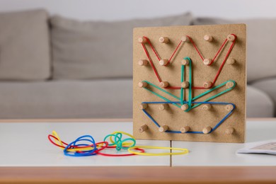 Wooden geoboard with flower made of rubber bands on white table in room, space for text. Motor skills development