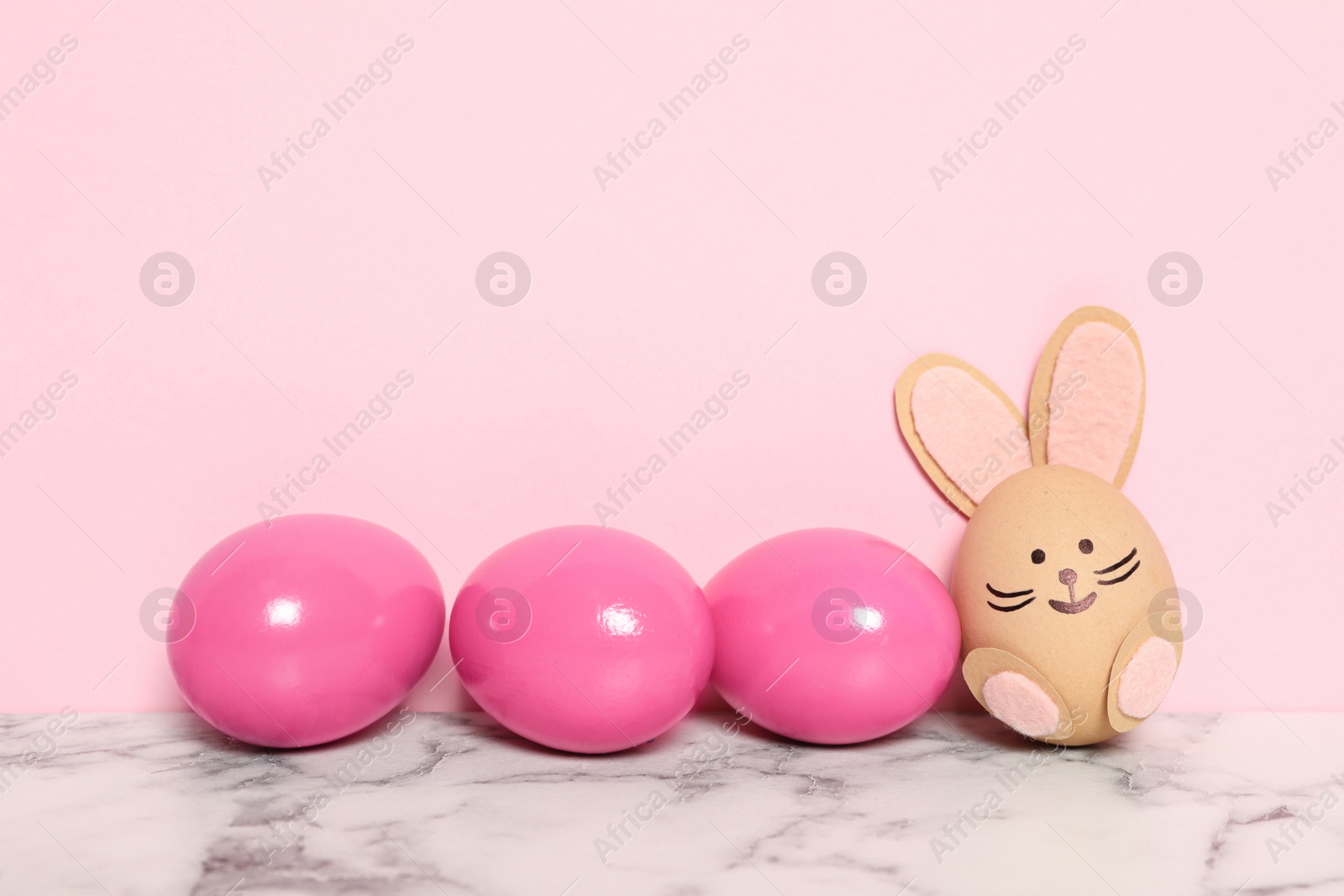 Photo of Bright Easter eggs and white one as cute bunny on marble table against pink background, space for text