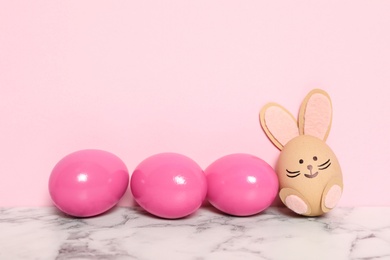 Bright Easter eggs and white one as cute bunny on marble table against pink background, space for text