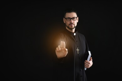 Image of Priest with Bible making blessing gesture and holy light on dark background, space for text