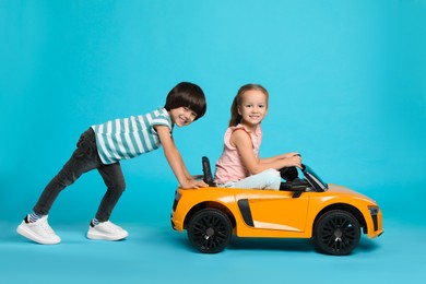 Photo of Cute boy pushing children's electric toy car with little girl on light blue background