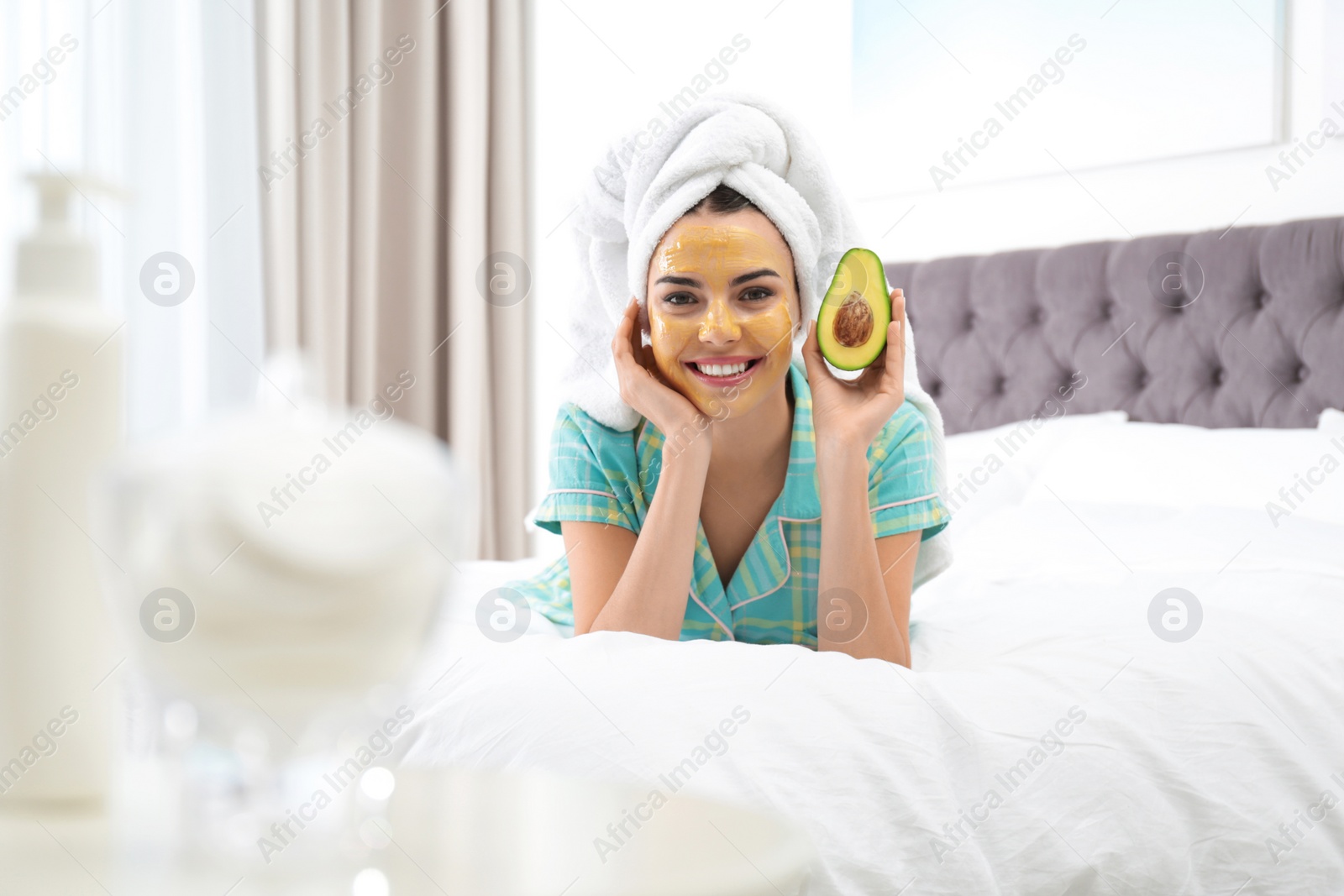 Photo of Young woman with cleansing mask on her face holding avocado in bedroom