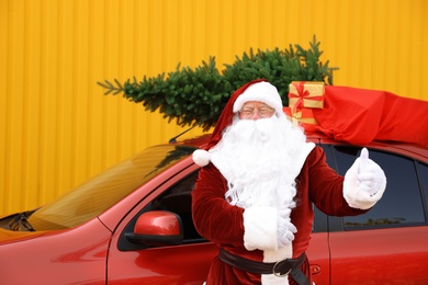 Photo of Authentic Santa Claus near car with fir tree and bag fullpresents on roof against yellow background