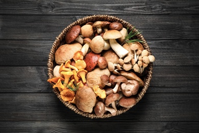 Photo of Different fresh wild mushrooms in wicker bowl on black wooden table, top view