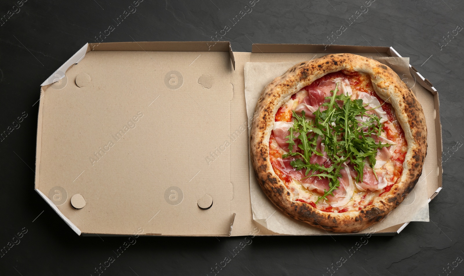 Photo of Tasty pizza with meat and arugula in cardboard box on black table, top view