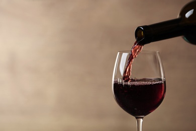 Pouring red wine into glass from bottle against blurred beige background, closeup. Space for text