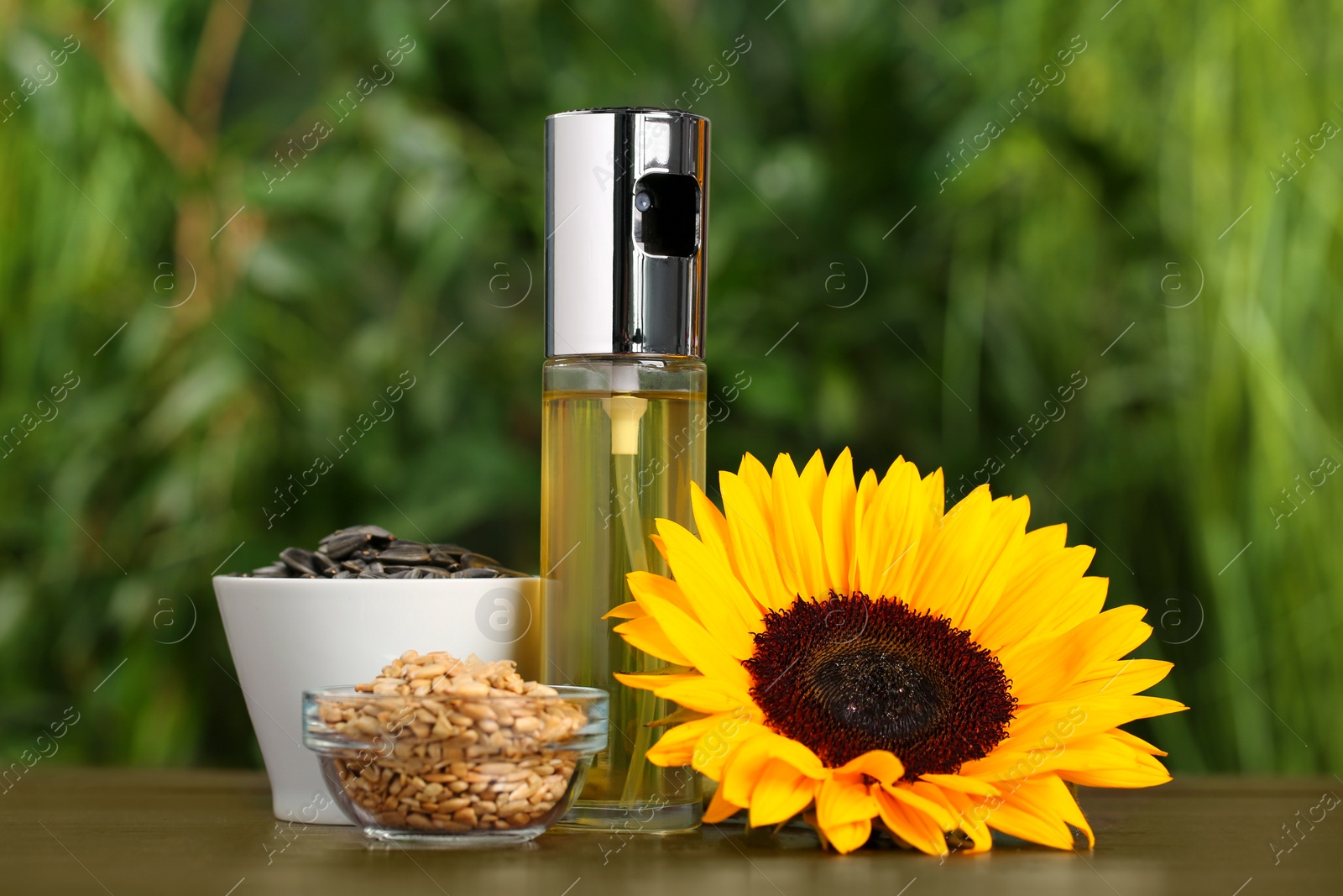 Photo of Spray bottle with cooking oil, sunflower seeds and flower on wooden table against blurred green background