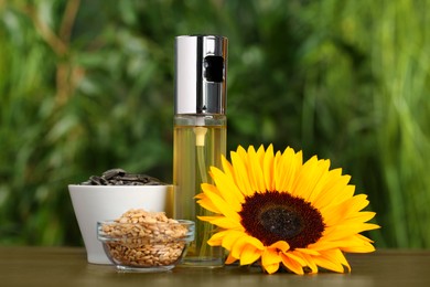 Photo of Spray bottle with cooking oil, sunflower seeds and flower on wooden table against blurred green background