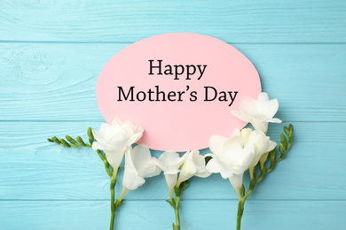 Image of Card with text Happy Mother's Day and beautiful flowers on light blue wooden background, flat lay